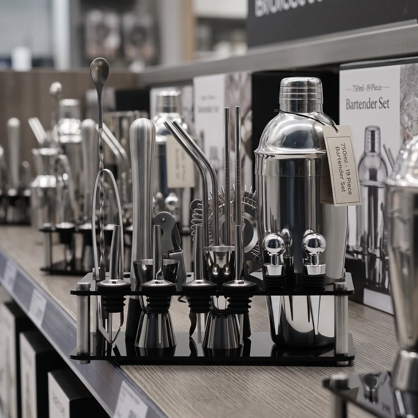 A 7-piece stainless steel bartender set displayed on a black stand on a retail shelf. The set includes a shaker, strainer, muddler, bar spoon, jigger, and various metal tools, with a small tag attached to the shaker.