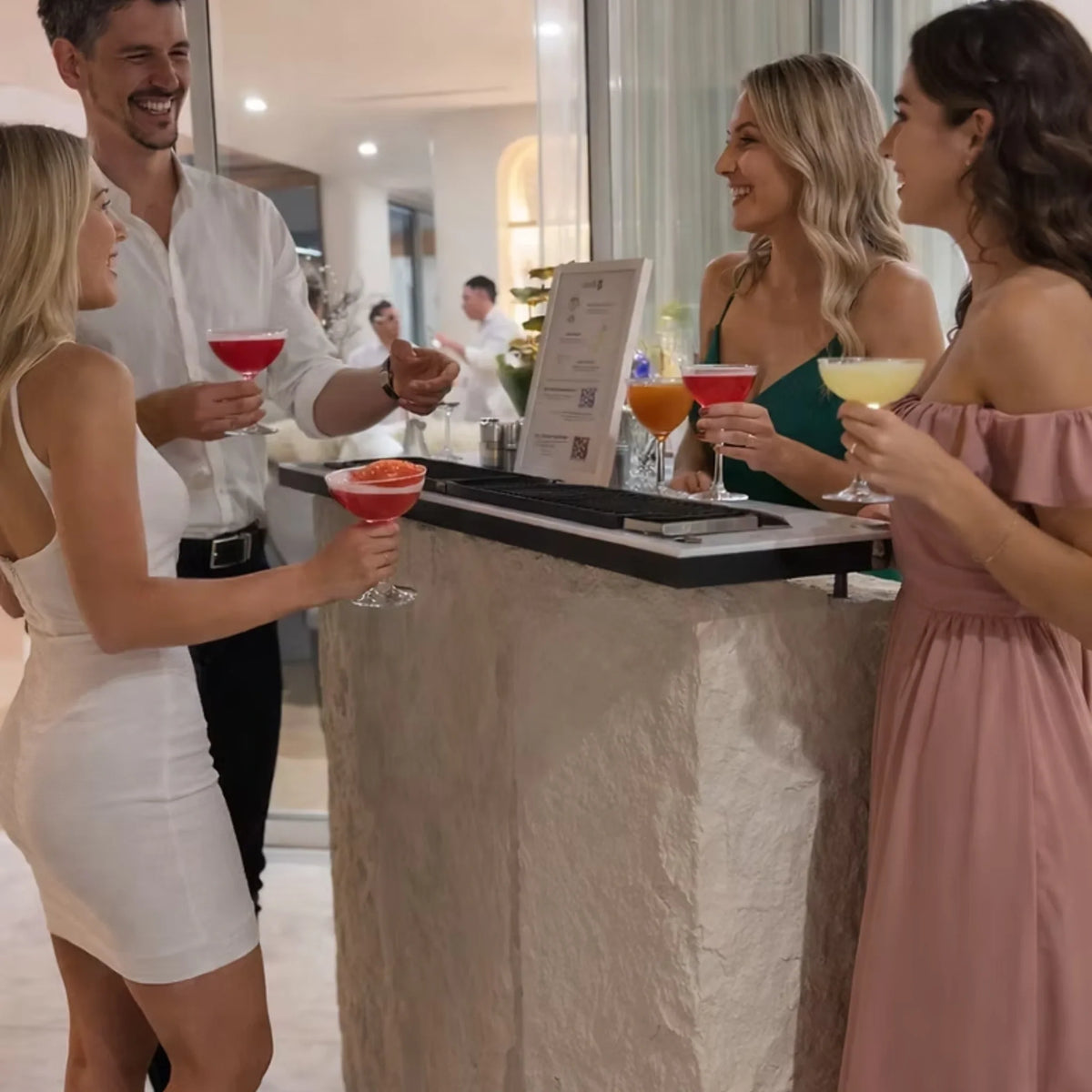 Four people gathered around a bar counter in a bright indoor venue, holding colorful cocktails. Two women in white and pink dresses stand on the left and right, smiling and talking with a man in a white shirt behind the counter.