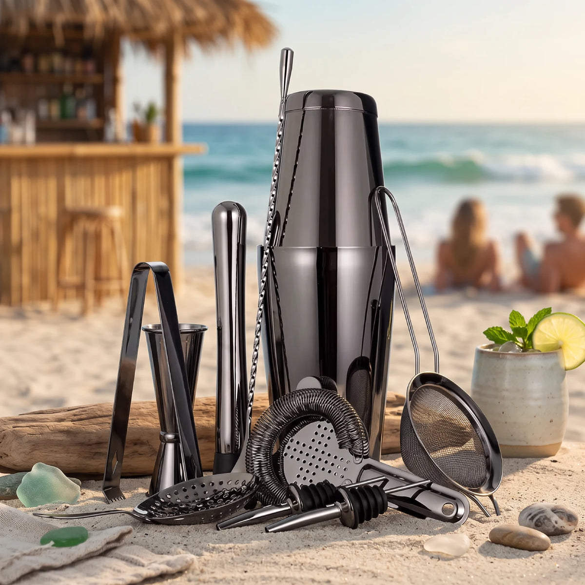 A black metal cocktail bar tool set arranged on a sandy beach with the ocean and a thatched beach bar blurred in the background. The set includes a shaker, strainer, muddler, jigger, and other bar accessories.