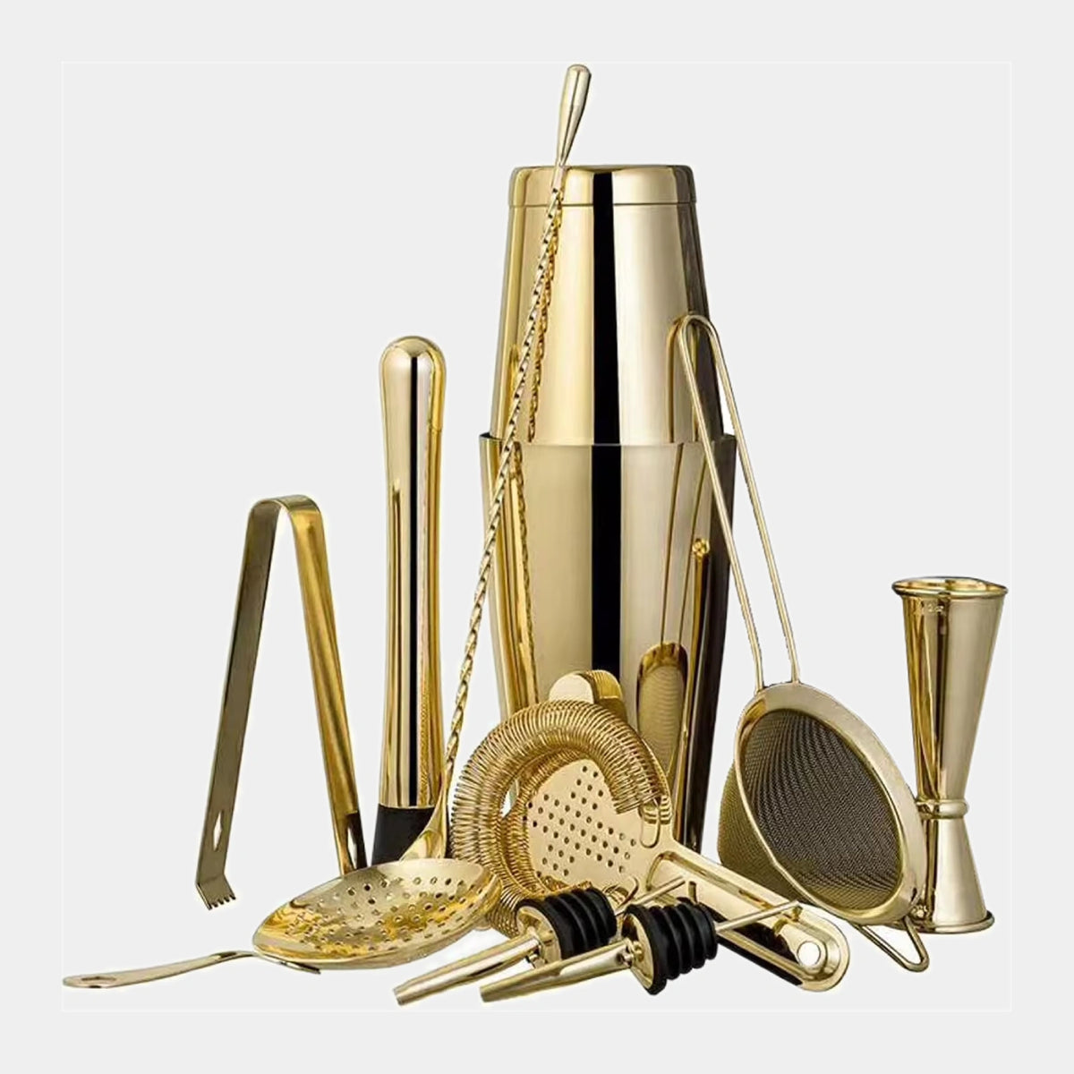 A gold-toned cocktail tool set arranged on a light gray background, including a shaker, strainer, jigger, muddler, spoon, and bar accessories.