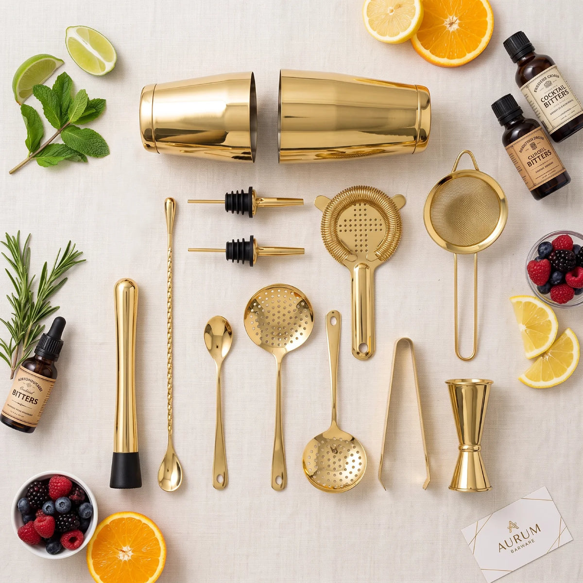 A gold-tone cocktail bar tool set arranged on a light fabric background with citrus slices, mint, rosemary, berries, and bitters bottles. The set includes shaker parts, strainers, spoons, tongs, a jigger, and bar accessories.