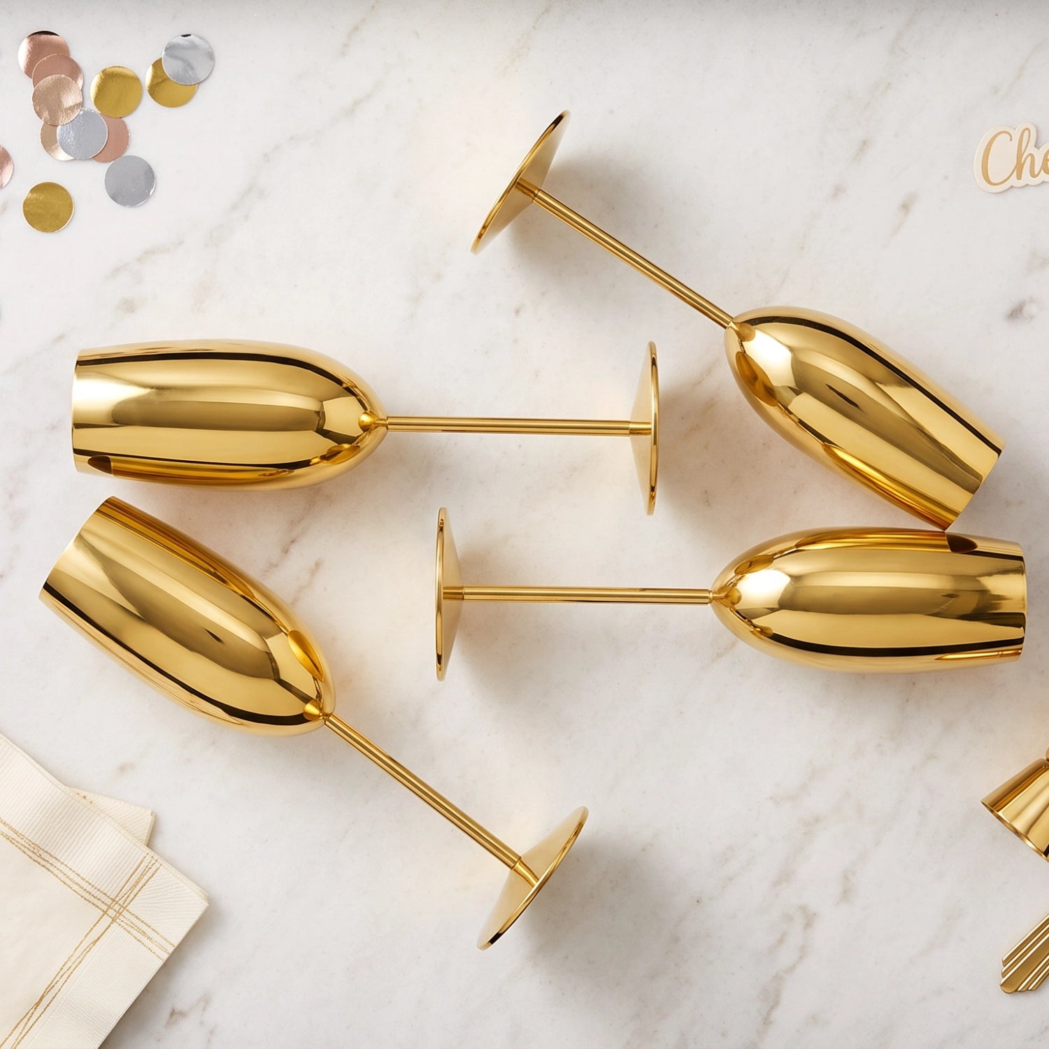 Four shiny gold champagne flutes arranged on a white marble surface with small confetti and napkins nearby.