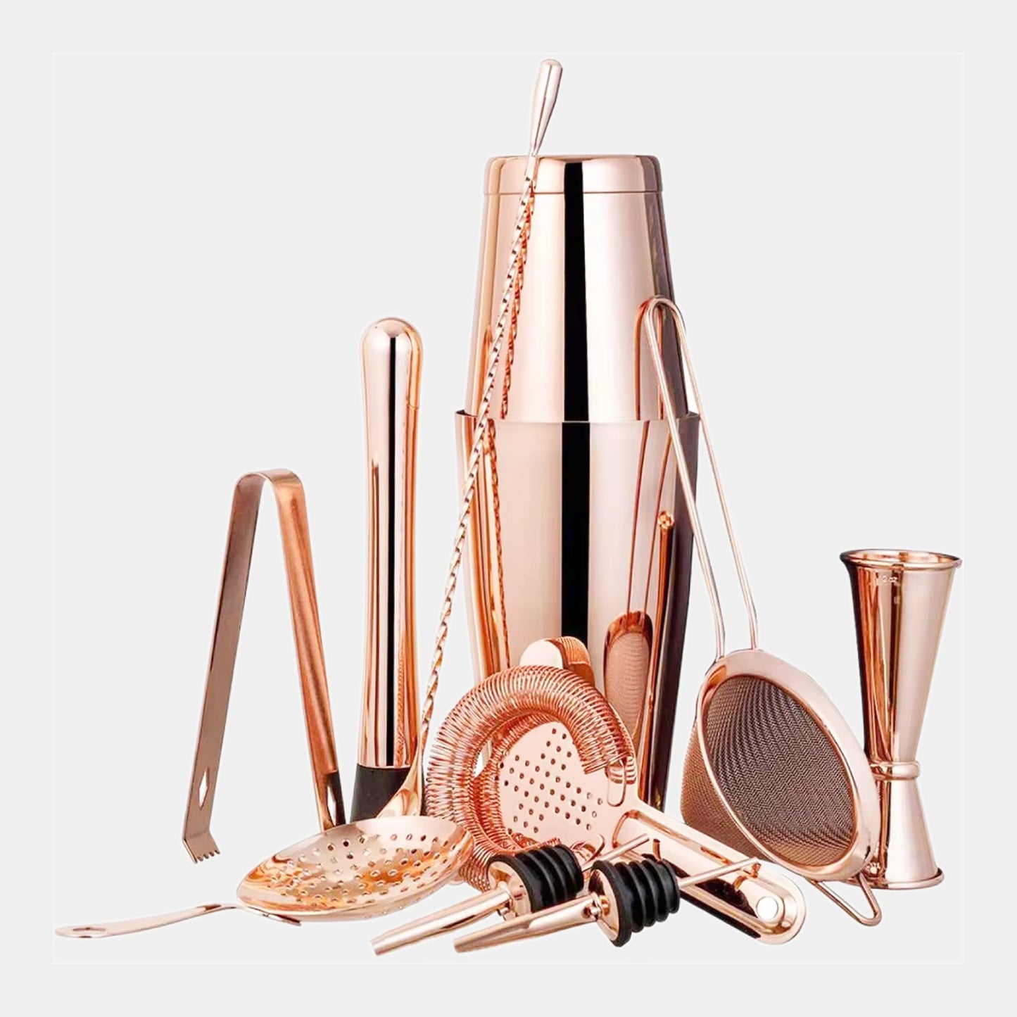 A rose gold cocktail bar tool set arranged on a light gray background, including a shaker, strainer, muddler, jigger, and other bartending accessories.