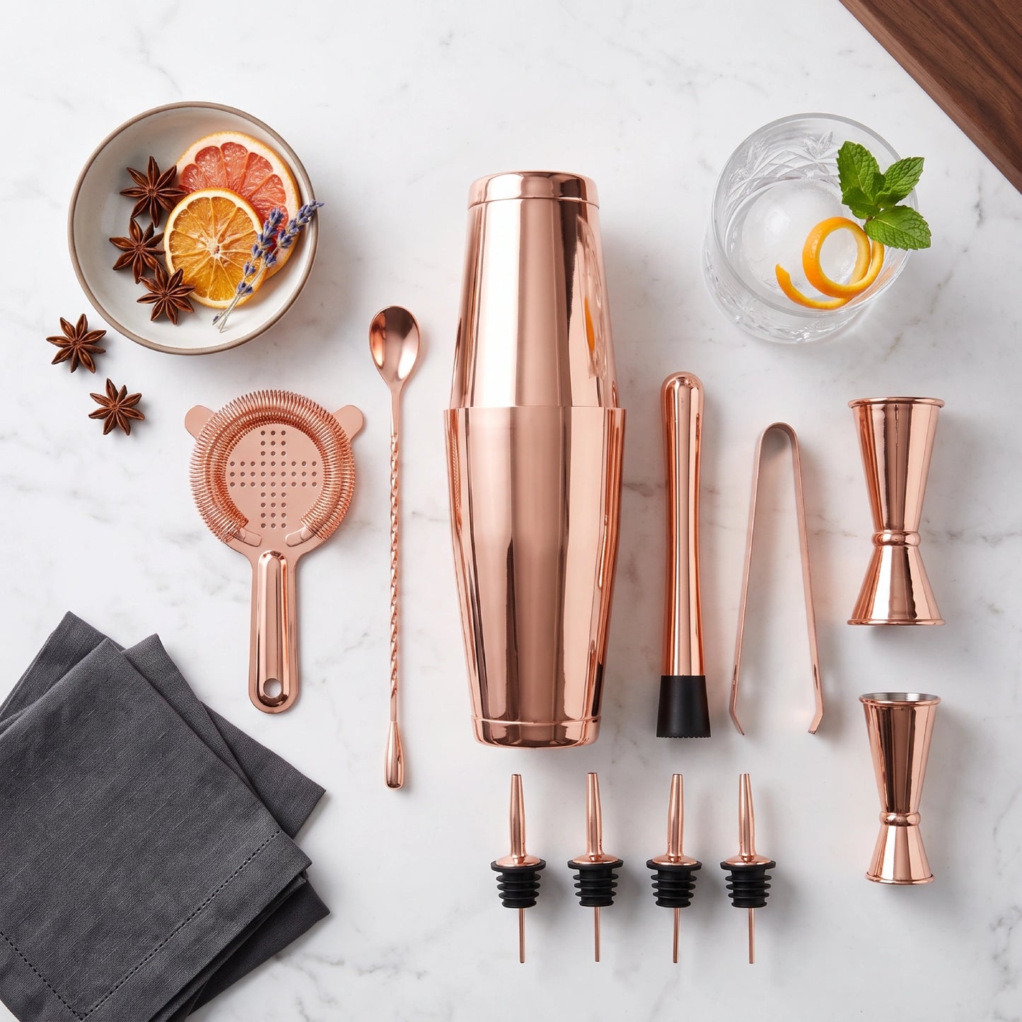 A copper cocktail bar tool set arranged on a white marble surface, including a shaker, strainer, spoon, jigger, ice tongs, pourers, and black stoppers, with garnish props and folded napkins around it.