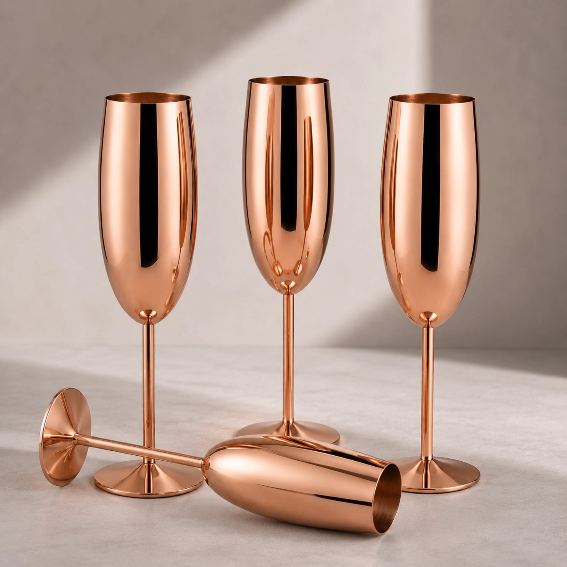 Three metallic champagne flutes with a polished copper or rose-gold finish, arranged on a light neutral surface against a soft beige background.