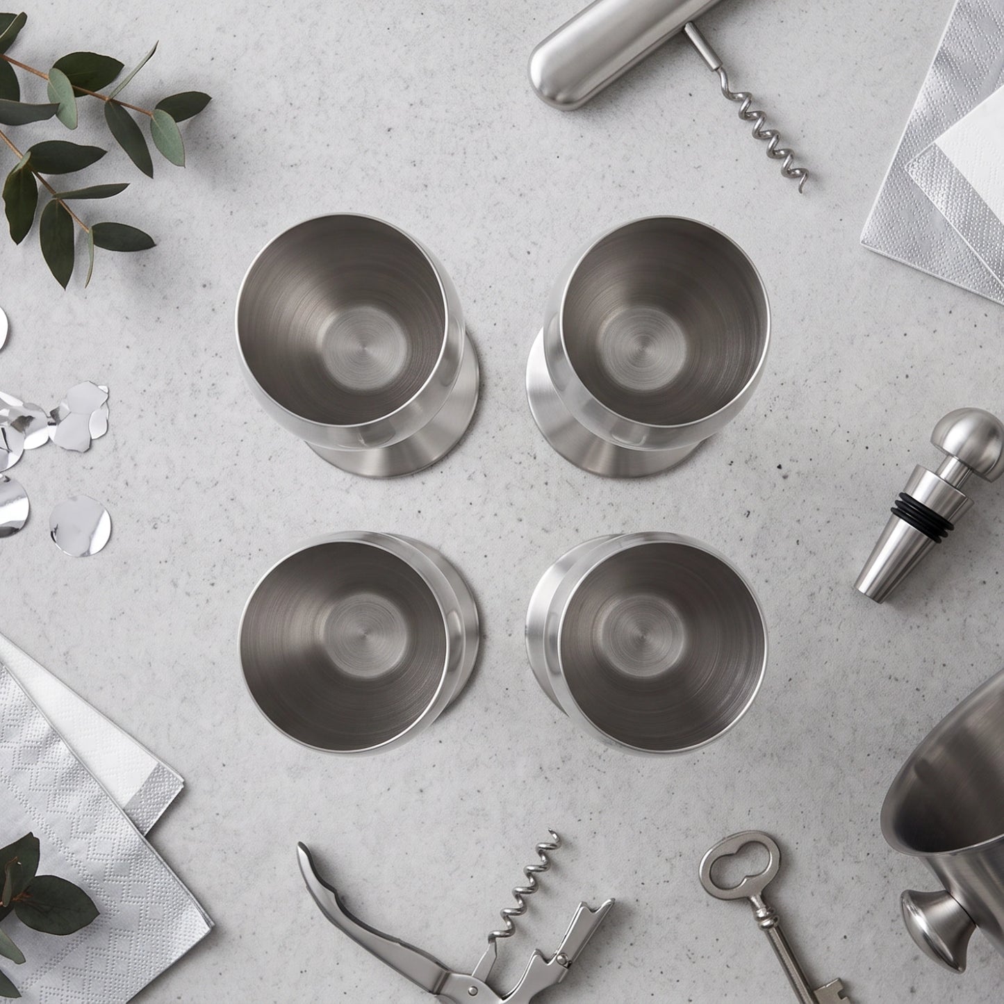 Four brushed stainless steel cocktail tumblers arranged in a 2x2 grid on a light gray stone surface, styled with bar tools, napkins, eucalyptus leaves, and confetti.