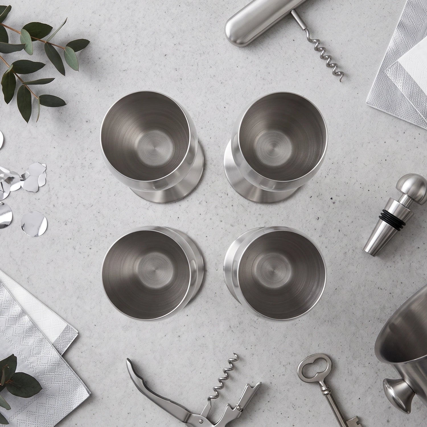 Four brushed stainless steel cocktail tumblers arranged in a 2x2 grid on a light gray stone surface, styled with bar tools, napkins, eucalyptus leaves, and confetti.