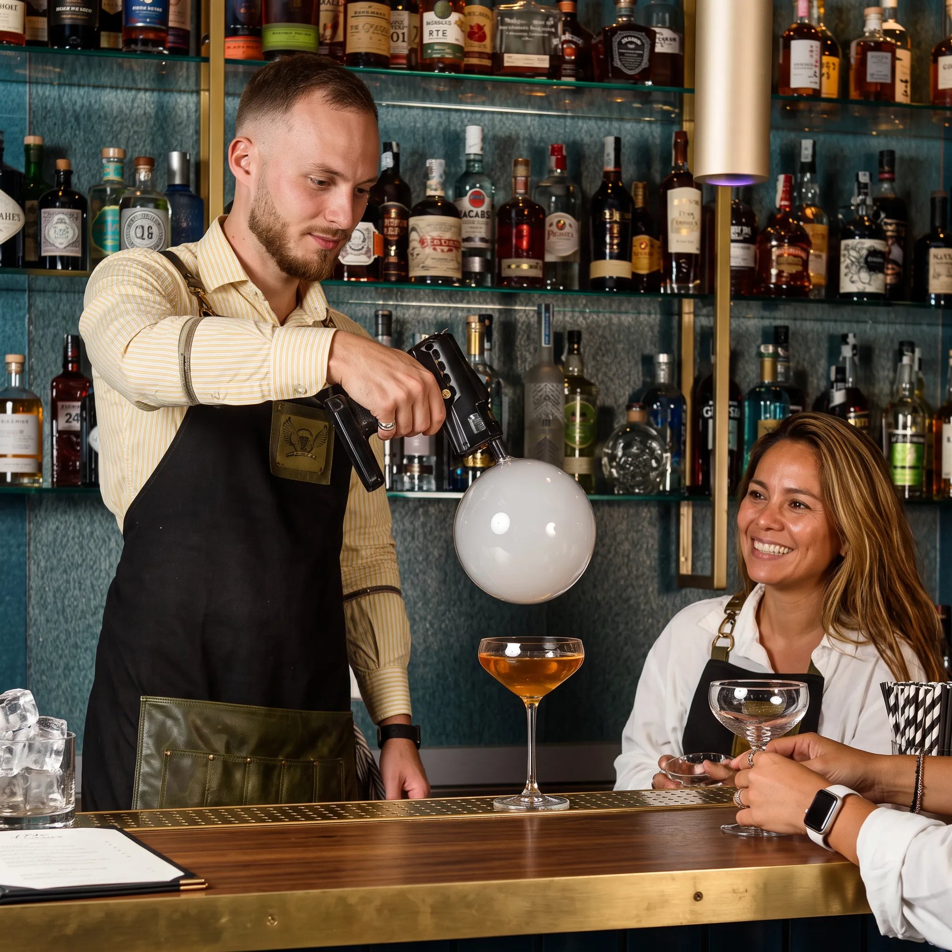 A bartender in a black apron and striped shirt uses a black handheld device to shoot a white balloon floating above a cocktail glass at a bar, with another person smiling in the background.