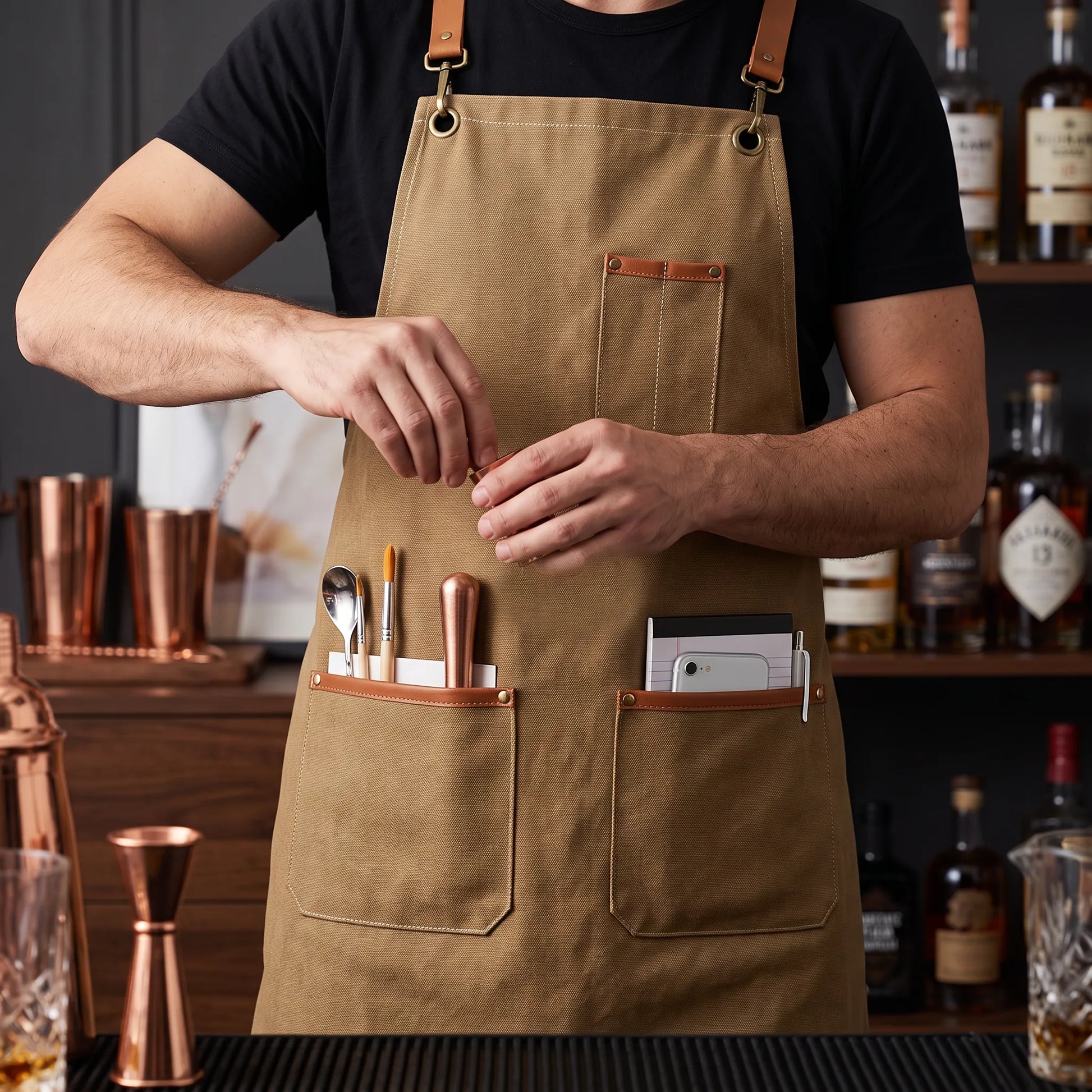 A man wearing a tan canvas apron with brown leather straps and pockets, holding bar tools in front of a bar counter with copper barware and bottles.