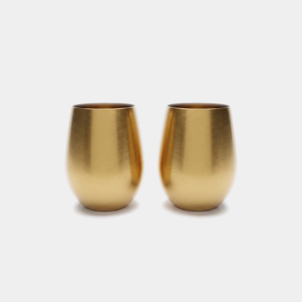 Two small metallic gold cups with a brushed finish, standing side by side on a light gray background.