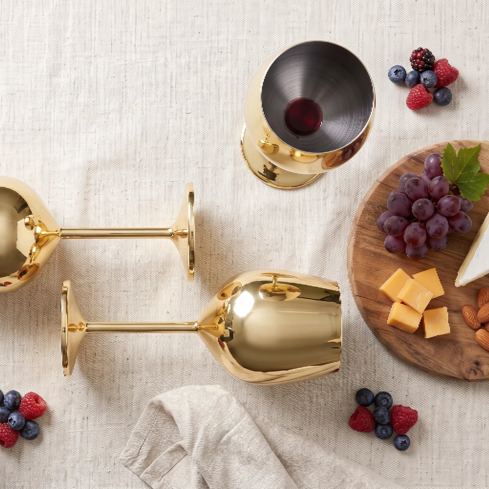 Two polished gold wine glasses arranged on a light beige fabric surface, with a wooden board of grapes, cheese, almonds, and berries nearby. No person or animal is present, and the scene is styled as a clean product flat-lay rather than a real environment, so subject_type is Product/Packaging.