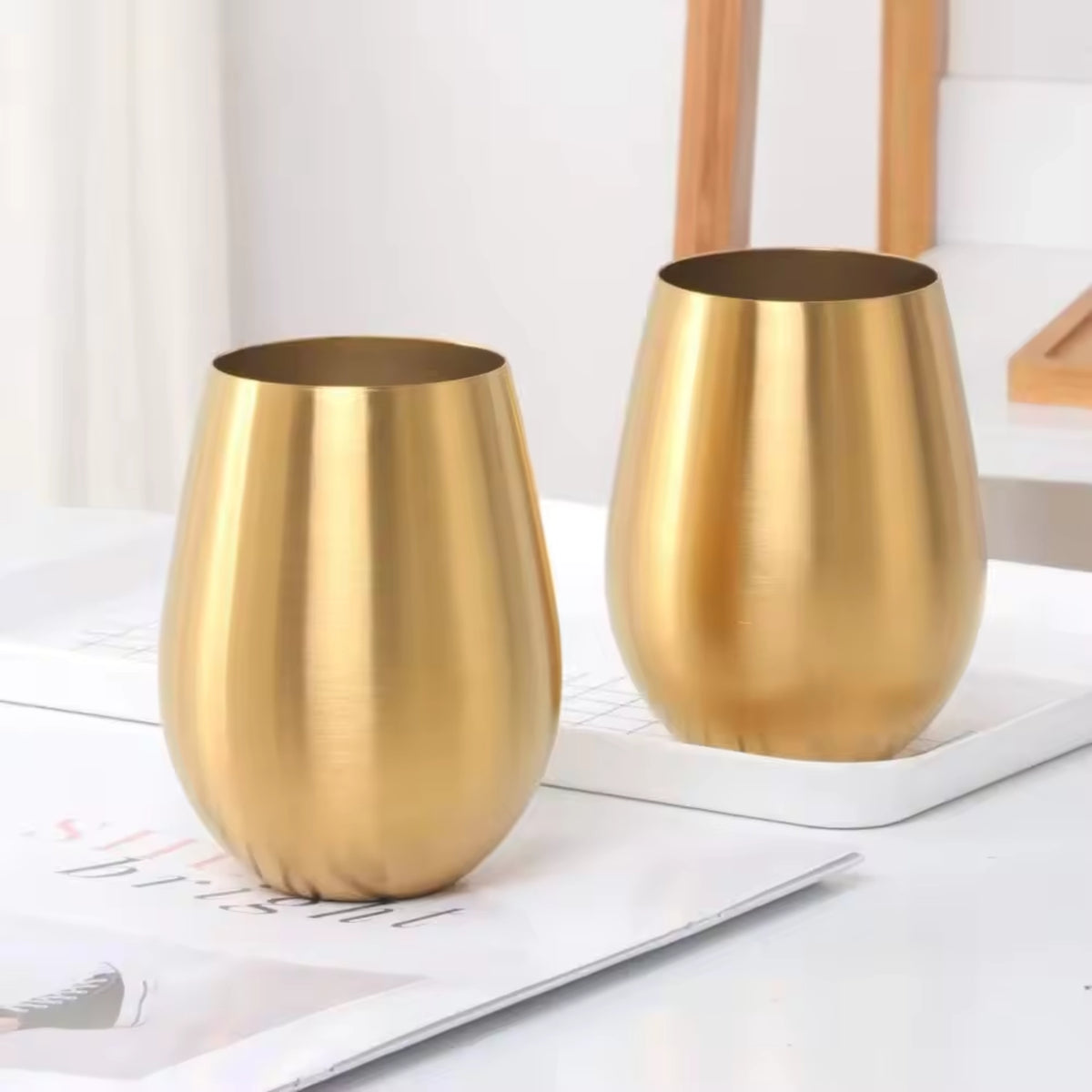 Two gold metal tumblers on a white surface with a light background