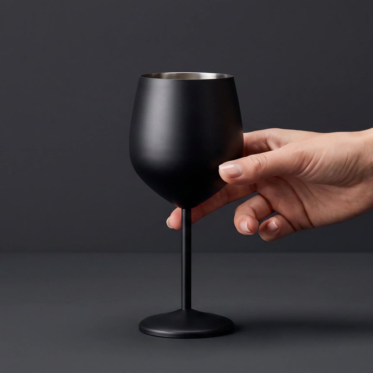 A matte black wine glass with a stem and circular base, held by a person's hand against a dark gray studio background.