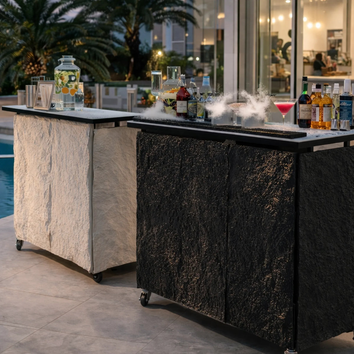 Two Premium Mobile Bars beside a pool at dusk, one with a light stone-textured cover and one with a dark charcoal cover.