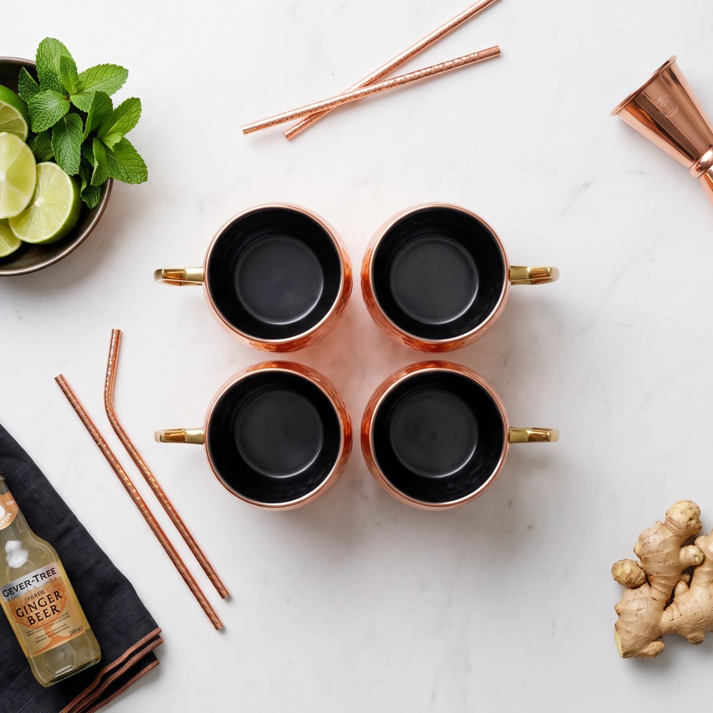 Four copper mugs with black interiors arranged in a 2x2 grid on a white marble surface, styled with copper straws, lime slices, mint, ginger, and a bottle of ginger beer.