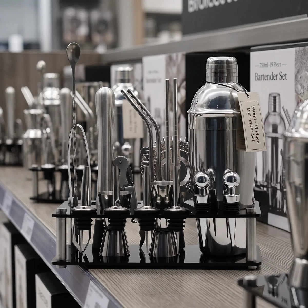 A 7-piece stainless steel bartender set displayed on a black stand on a retail shelf. The set includes a shaker, strainer, muddler, bar spoon, jigger, and various metal tools, with a small tag attached to the shaker.