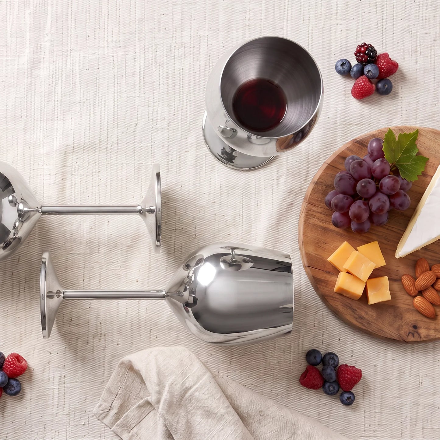Three polished stainless steel wine glasses arranged on a light beige textured tablecloth with food props including grapes, cheese, almonds, berries, and a folded napkin.