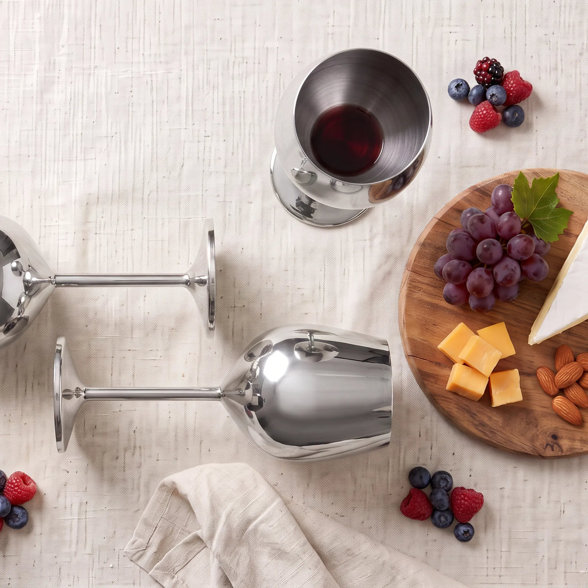 Three polished stainless steel wine glasses arranged on a light beige textured tablecloth with food props including grapes, cheese, almonds, berries, and a folded napkin.