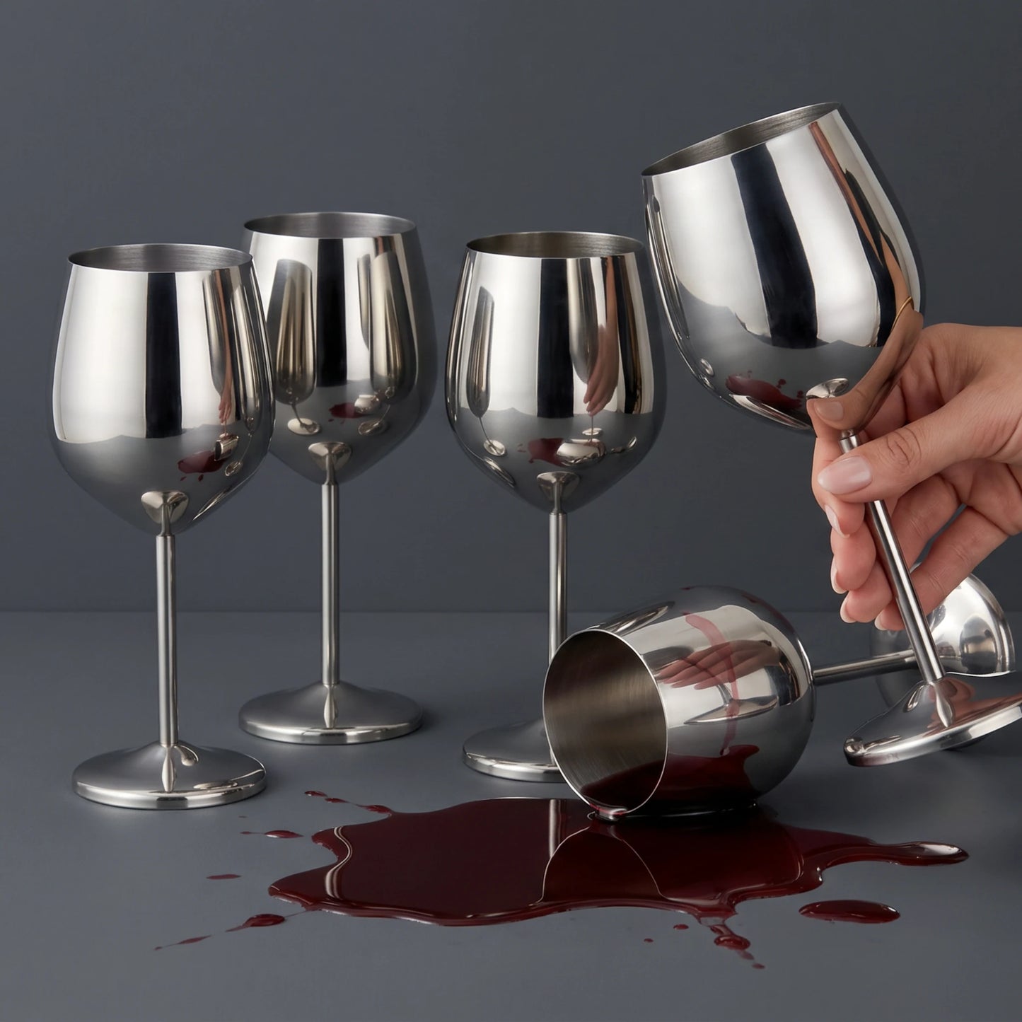 Four stainless steel wine glasses on a dark gray surface, with one glass tipped over spilling a dark red liquid and a hand holding another glass.