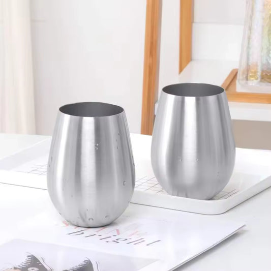 Two brushed stainless steel stemless wine glasses with a water droplet finish, resting on a white tray on a white table with a magazine underneath.