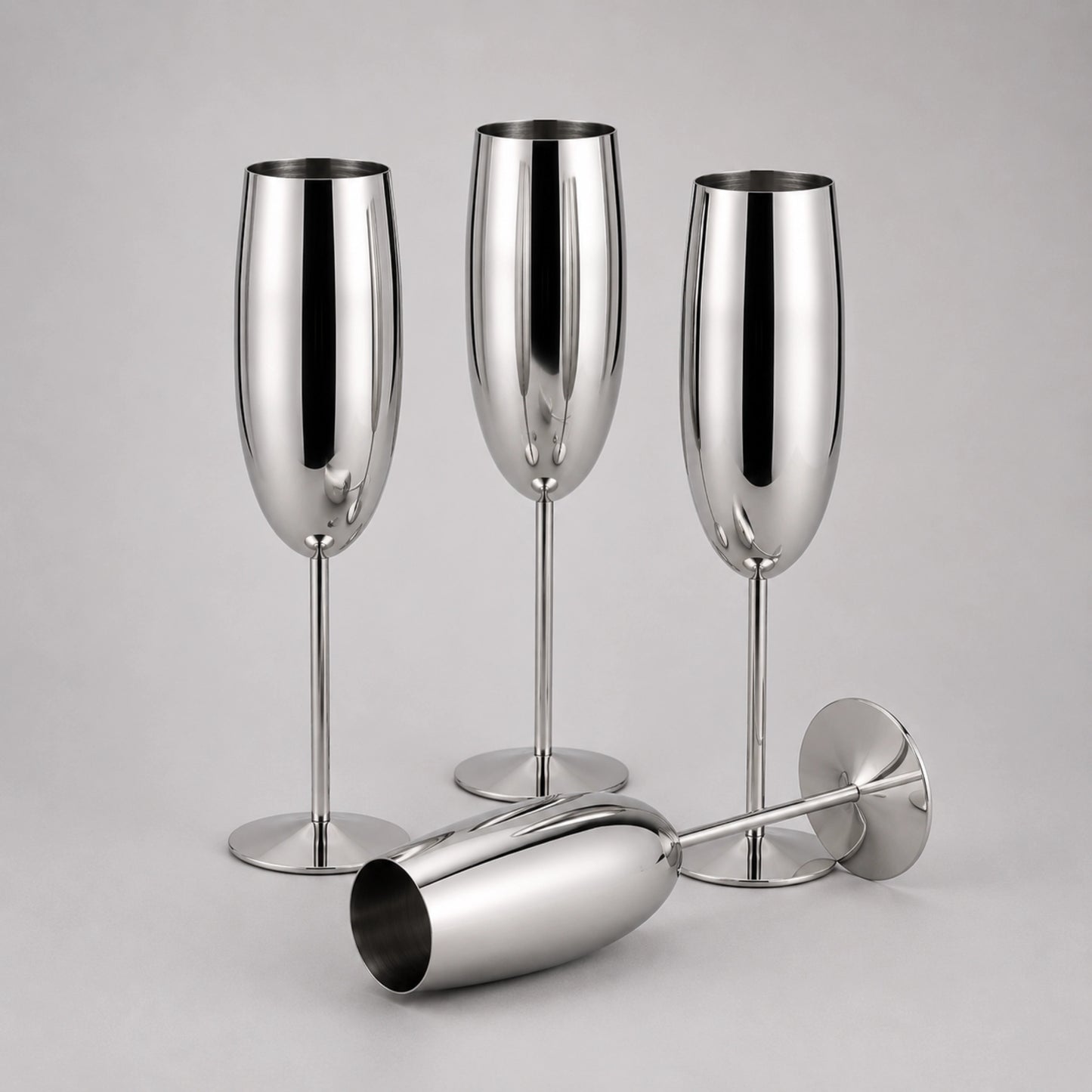 Three polished stainless steel champagne flutes with stems and bases, plus one matching coupe-shaped glass lying on its side, photographed on a light gray studio background.