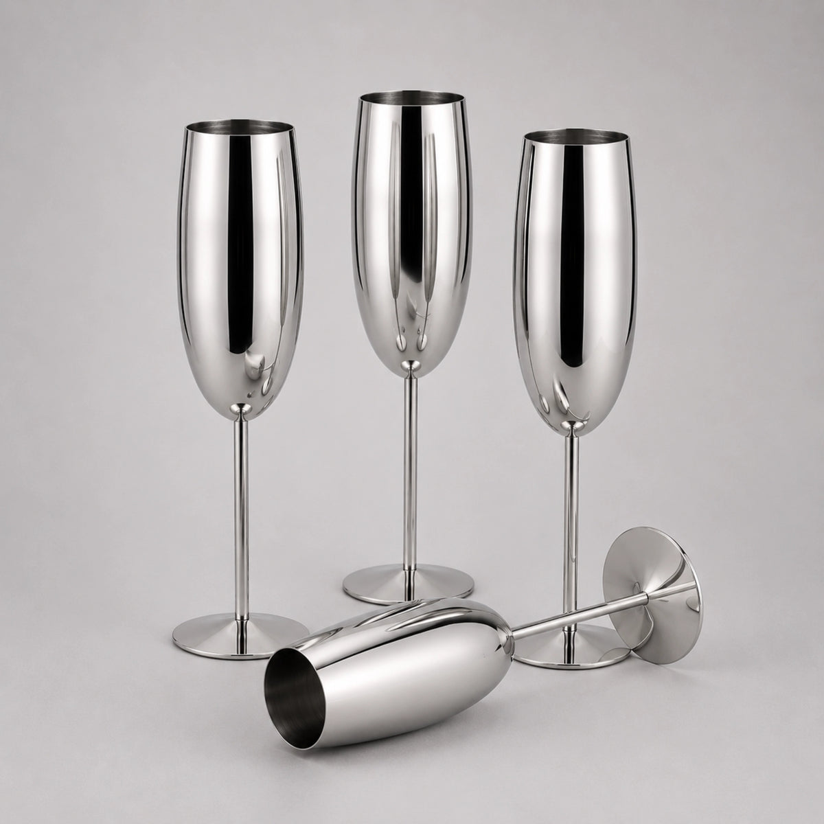 Three polished stainless steel champagne flutes with stems and bases, plus one matching coupe-shaped glass lying on its side, photographed on a light gray studio background.
