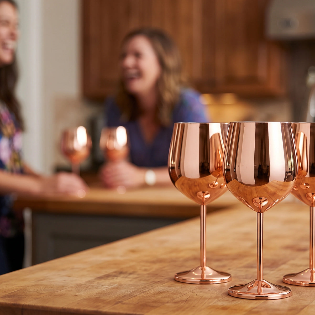 Three shiny copper-colored wine glasses on a wooden countertop in a kitchen, with two blurred people holding matching glasses in the background.